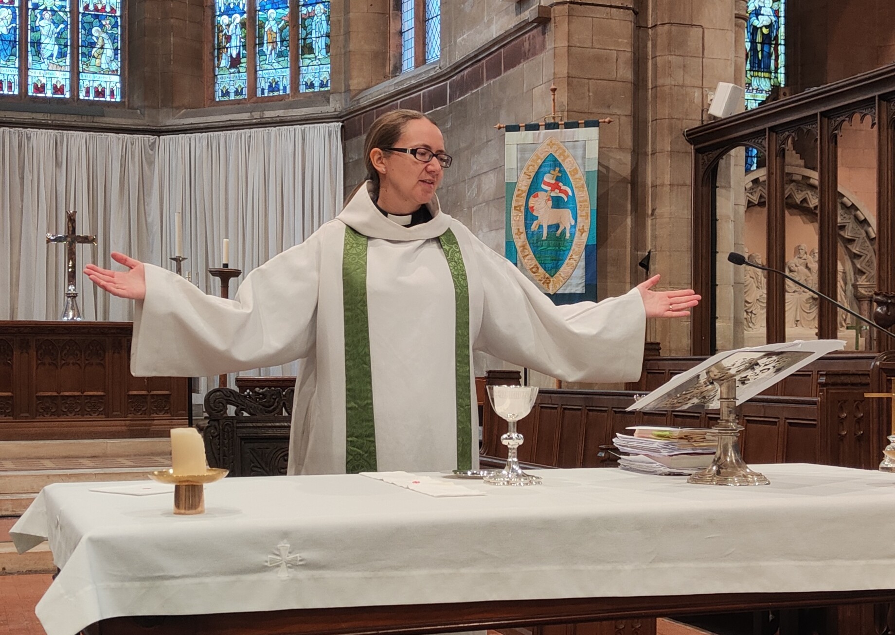A female priest is standing with arms outstretched behind the altar, a chalice and patten are on the altar