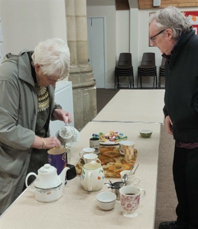 A lady pouring a drink, a table with cups and chocolate biscuits, a man asking for a drink